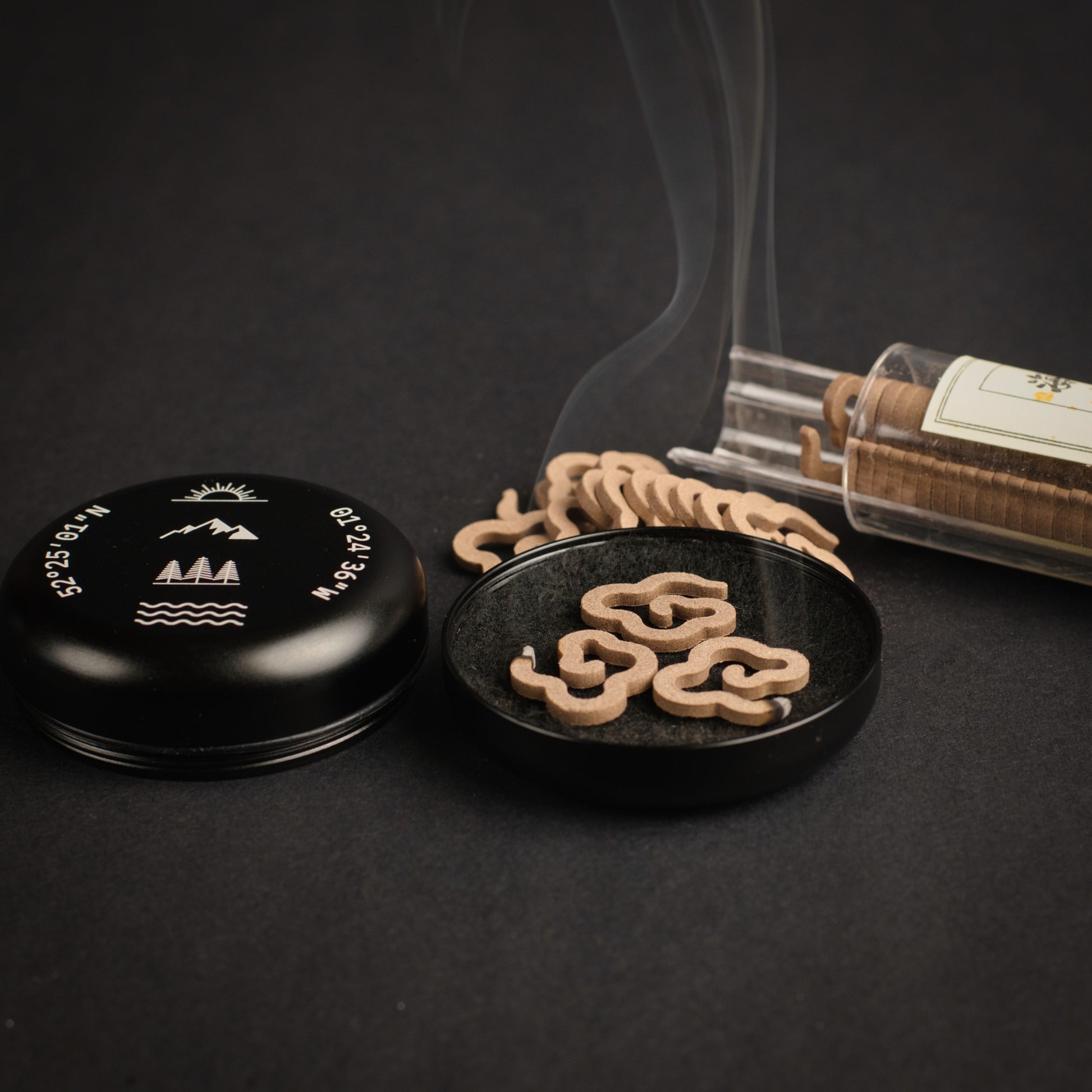 Portable incense burner in black with engraved coordinates and nature-inspired symbols, shown burning agarwood cloud incense on a carbon felt insert