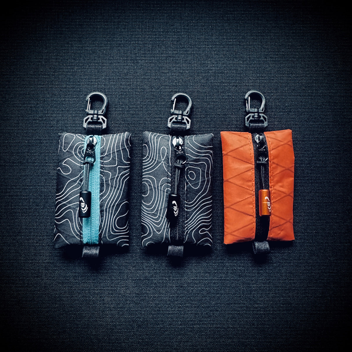 Three X-Pac micro utility pouches in topo black, topo blue and orange, each with YKK zip, engraved Tread Lite pull and swivel carabiner hook.