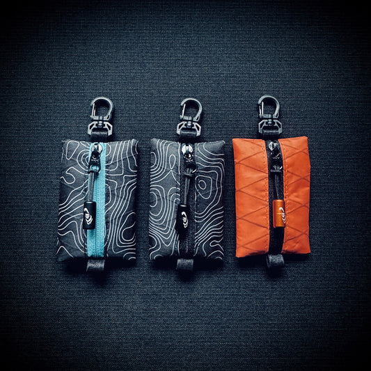 Three X-Pac micro utility pouches in topo black, topo blue and orange, each with YKK zip, engraved Tread Lite pull and swivel carabiner hook.