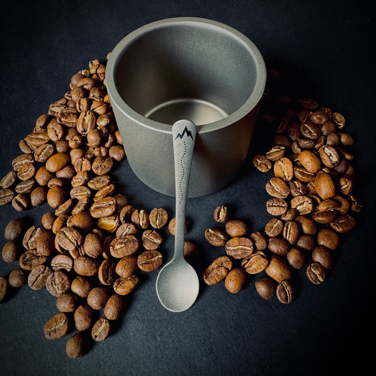 Grade 5 titanium coffee spoon with Trail mountain and dotted path design, displayed with coffee beans and a titanium cup.