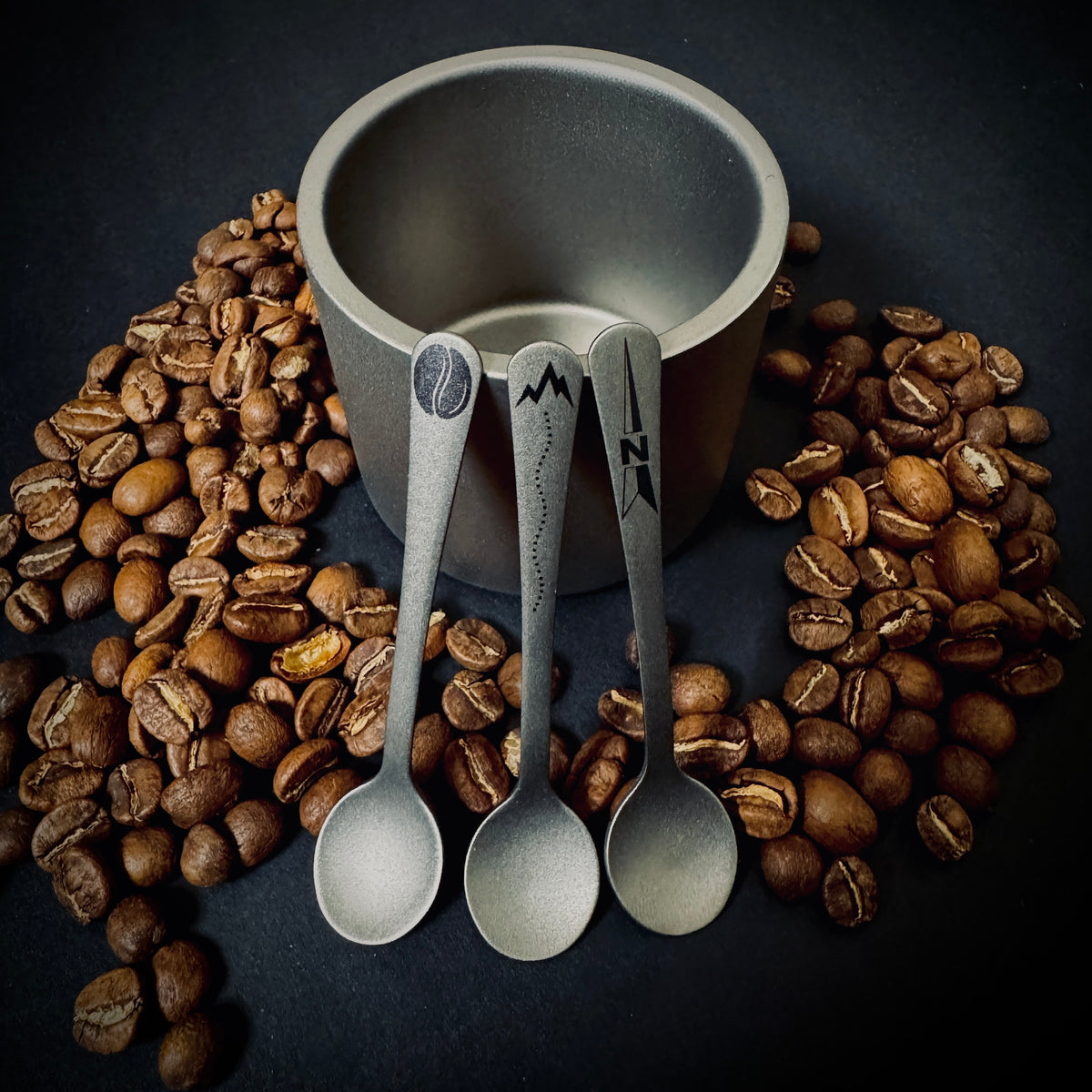 Three Grade 5 titanium coffee spoons with Coffee, Trail and North laser marked designs, displayed with coffee beans and a titanium cup.