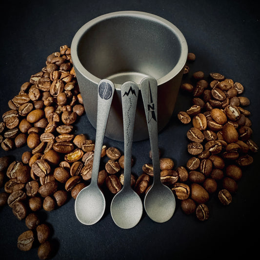 Three Grade 5 titanium coffee spoons with Coffee, Trail and North laser marked designs, displayed with coffee beans and a titanium cup.