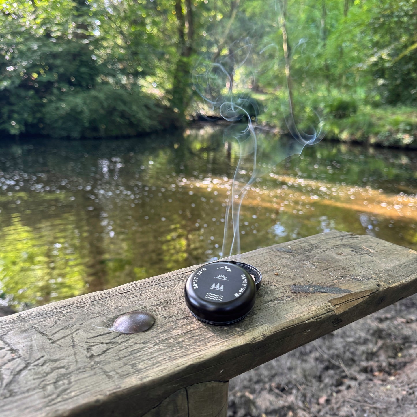Portable incense burner beside a woodland river with incense smoke rising, personalised with engraved coordinates and outdoor-inspired glyphs.